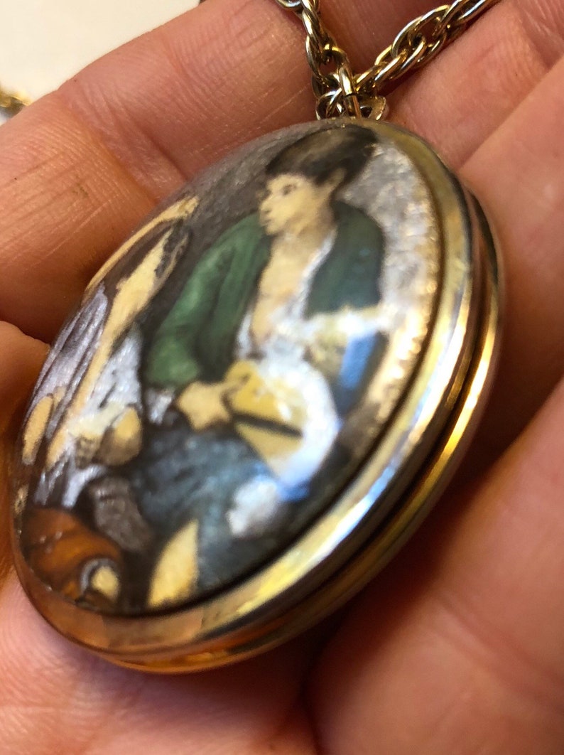 May include: Oval pendant necklace with a gold-tone chain. The pendant features a miniature painting of a person wearing a green jacket and white shirt. The painting has a gold-tone border.