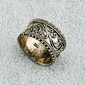 May include: A silver ring with an intricate, ornate design featuring swirling patterns. The ring is wide and has a rounded band.