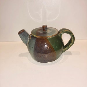 May include: A small, brown, green, and blue ceramic teapot with a lid. The teapot has a rounded body and a curved handle.