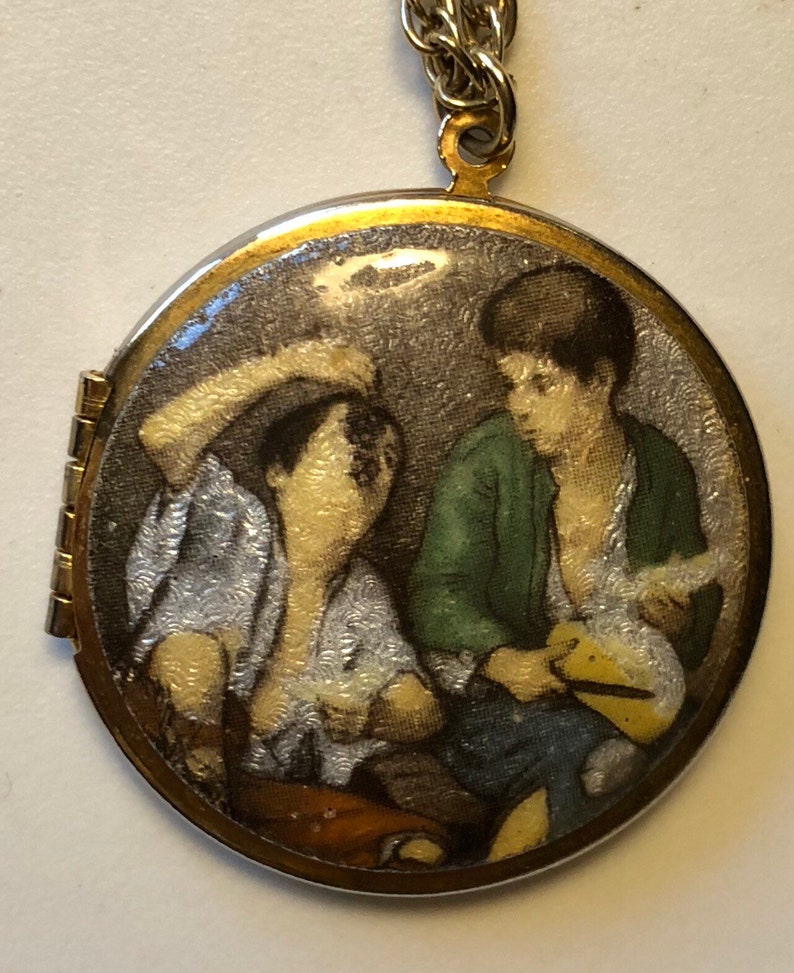 May include: A round, hinged pendant necklace with a gold-tone frame and a silver chain. The pendant features a painted scene of two figures in a room, with one wearing a green jacket. The pendant is approximately 1.5 inches in diameter.