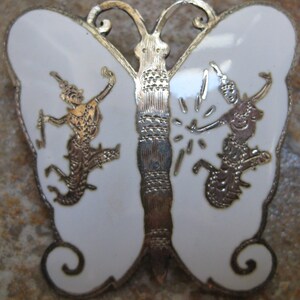 May include: A white enamel butterfly brooch with silver filigree detailing. The butterfly has two figures on each wing, each figure is in a dancing pose.