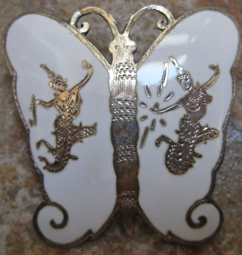 May include: A white enamel butterfly brooch with silver filigree detailing. The butterfly has two figures on each wing, each figure is in a dancing pose.