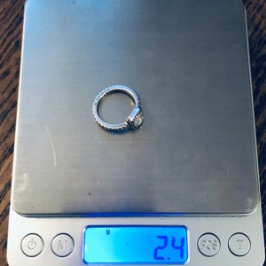 May include: A silver ring with a clear stone on a digital scale that reads 2.4 grams.