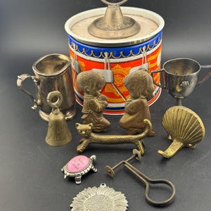 May include: A collection of vintage metal objects, including a small silver cup, a bell, a drum-shaped container, and a turtle-shaped trinket with the words "Keep it simple." Other items include a cat-shaped object and a sunflower pendant.