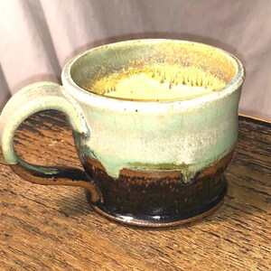 May include: A handmade ceramic mug with a green, brown, and yellow glaze. The mug has a rounded body and a wide handle.
