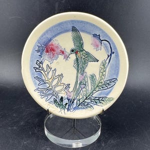 以下が含まれることがあります： 緑と赤色のハチドリが飛んでいる様子を描いた陶器の皿。ピンクと紫の花の近くにいます。 皿の中央はクリーム色で、青い縁と緑の葉があります。