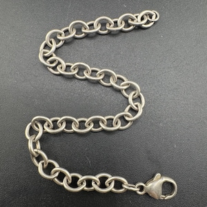 May include: A silver-colored chain bracelet with a lobster clasp. The bracelet is made of oval-shaped links and is displayed on a dark surface. The chain is a simple, elegant design.