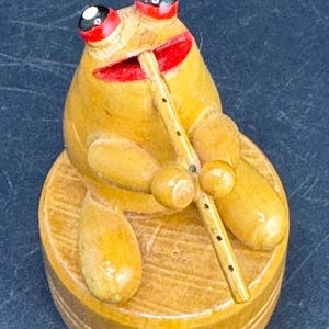 May include: A small, light brown wooden figurine of a frog sitting on a circular wooden base. The frog has prominent red eyes with black and white pupils, a red mouth, and is holding a thin, light-colored wooden flute to its lips.