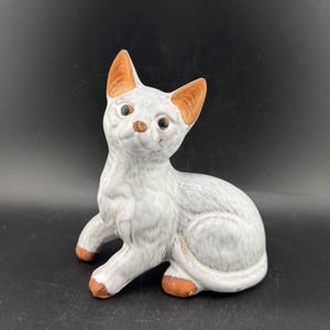 May include: A ceramic cat figurine in a seated pose. The cat is primarily white with brown accents on the ears, nose, and paws. The figurine has a textured surface and detailed facial features, including dark eyes.