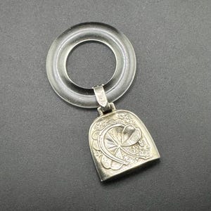 May include: A clear, circular teething ring with a silver bell pendant. The bell features an ornate floral design and star accents. The ring is made of a transparent material, and the bell is made of silver.