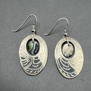 May include: Silver-coloured drop earrings with an oval, engraved design. Each earring features a central, oval-shaped abalone shell inlay. The earrings have a hook closure.