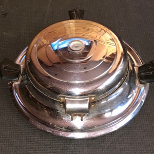 Könnte beinhalten: Ein vintage Waffeleisen aus Chrom mit schwarzen Kunststoffgriffen. Das Waffeleisen hat ein kreisförmiges Design mit einer erhabenen Mitte und einer strukturierten Oberfläche.