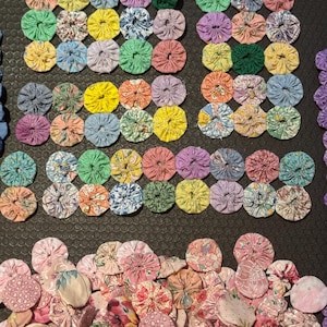 Puede incluir: Una colección de yo-yos de tela coloridos en varios tamaños y patrones. Los yo-yos están dispuestos en filas y dispersos, mostrando una variedad de telas florales y estampadas en tonos de rosa, amarillo, verde y azul. El fondo es oscuro.