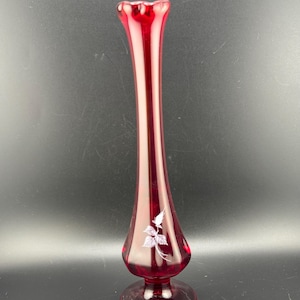 Puede incluir: Un jarrón de vidrio rojo, alto y delgado, con un diseño floral blanco. El jarrón tiene una base acampanada y una abertura estrecha.
