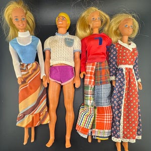 Op de afbeelding: Vier vintage poppen worden getoond. Eén pop draagt een blauw en wit topje met een gestreepte rok. Een andere pop draagt een witte trui en een paarse short. De derde pop draagt een rode top en een geruite broek. De laatste pop draagt een bloemenjurk.