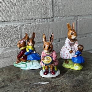 May include: Four ceramic figurines of rabbits, each with a different pose and outfit. One rabbit is playing a drum, another is riding a sled, and two are standing. The figurines are painted in bright colors and have detailed features.