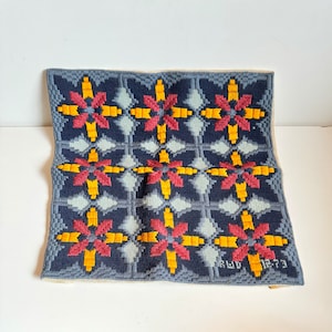 Peut inclure: Un coussin en point de croix bleu et blanc avec un motif géométrique de fleurs jaunes et rouges. Le coussin porte la date "18 juillet 1973" brodée dans le coin inférieur droit.