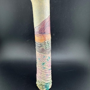 Op de afbeelding: Een lange, cilindrische keramische vaas met een textuuroppervlak en een verscheidenheid aan kleuren. De vaas heeft kleurstroken in beige, paars, oranje, grijs en roze, met een turquoise basis. De vaas is ongeveer 30 cm hoog.