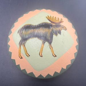 May include: A round, hand-painted wooden decorative piece featuring a moose illustration. The moose is depicted in shades of brown, black, and yellow, set against a light green background within an orange zig-zag border. The outer edge is dark green.