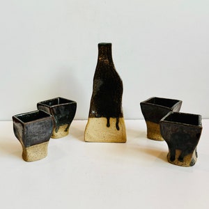 May include: Set of four ceramic sake cups and a sake bottle. The cups and bottle are all black with a brown glaze and have a white base.