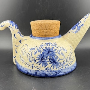 May include: A ceramic oil cruet with a cork stopper. The cruet is cream-colored with blue floral and scrollwork designs. It has a curved handle and a spout. The cork stopper is a light brown color.