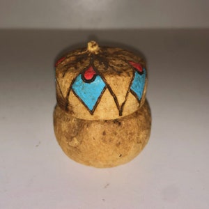 May include: A small, round, brown ceramic container with a lid. The lid is decorated with a geometric pattern of blue and red diamonds.