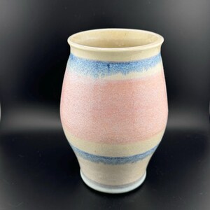 May include: A ceramic vase with a light pink, cream, and blue striped design. The vase has a rounded, slightly flared shape.