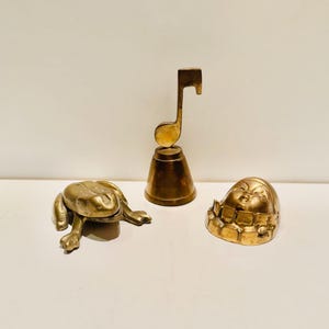 Pode incluir: Três pequenos sinos de latão, um com a forma de um sapo, outro com uma nota musical e outro com um rosto.