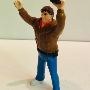 May include: A plastic action figure of a man wearing a brown jacket, blue jeans, and white sneakers. He is holding a black object in his right hand and has his left arm raised.