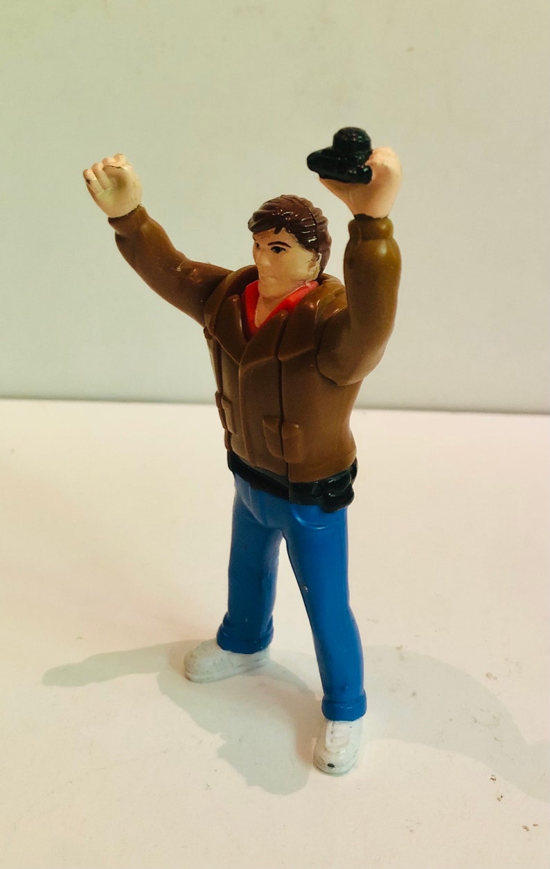 May include: A plastic action figure of a man wearing a brown jacket, blue jeans, and white sneakers. He is holding a black object in his right hand and has his left arm raised.
