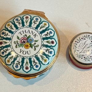 May include: Two round, decorative trinket boxes with floral designs. The larger box has a white background with a green floral border and the words "Thank You" in the center. The smaller box has a white background with a green floral border and the words "I Love You" in the center.