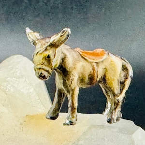 May include: A small, hand-painted donkey figurine with a saddle. The donkey is primarily tan and brown, with a white muzzle and ears. The saddle is orange. The figurine is standing on a white, crystalline surface against a dark background.