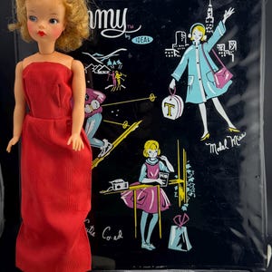 May include: A vintage Tammy doll wearing a red strapless dress and red shoes, standing on a cream-colored stand. Behind her is a black carrying case with illustrations of a woman in a blue coat and other retro graphics. The case is labeled "Tammy by Ideal."