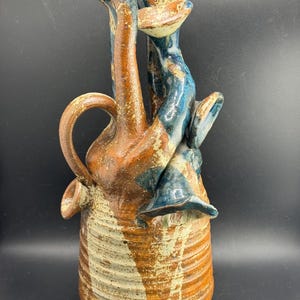 May include: A ceramic vessel with a ribbed base, featuring a mottled brown and cream glaze. The top section has multiple elongated, tubular spouts and flared openings, accented with deep blue and brown glaze drips and textured surfaces.