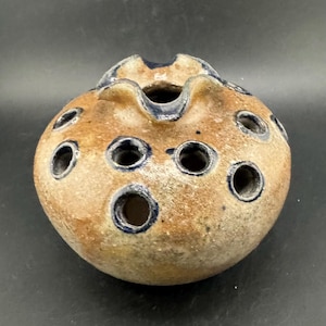May include: A round, ceramic flower frog with a textured, speckled brown surface. It features multiple circular holes with dark blue rims, and a ruffled opening at the top. The flower frog is designed to hold flowers in place.