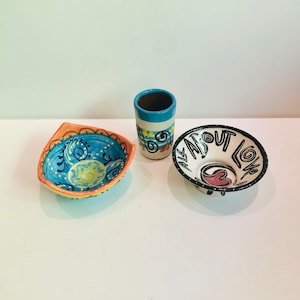 Pode incluir: Três tigelas de cerâmica, uma azul e branca com um design de espiral, uma branca com letras pretas que dizem "All About Love" e um coração rosa, e uma azul e branca com um design de espiral.