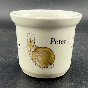 May include: Cream-colored ceramic cup featuring a brown and tan rabbit illustration. The text "Peter sat" is printed on the cup. The cup has a rounded shape with a slightly flared rim, suitable for holding liquids.