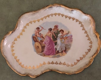 May include: A white porcelain serving tray with a gold rim and a painted scene of four figures in a chariot. The tray has a scalloped edge and a gold floral design.