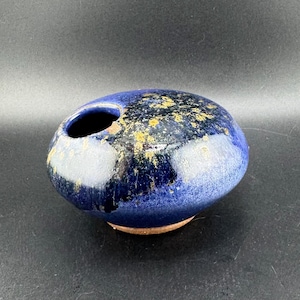 May include: A small, blue ceramic vase with a speckled glaze and gold flecks. The vase has a round, oval shape with a single opening at the top.
