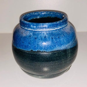 May include: A small, round ceramic vase. The upper half is a bright blue, fading into a darker, near-black, tone on the lower half. The vase has a wide opening and a slightly textured surface. A decorative item.