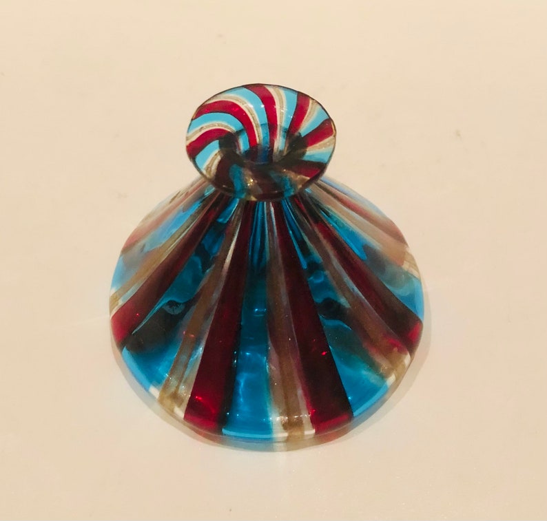 May include: A decorative glass bottle stopper with a spiral top. The stopper has alternating red, blue, and gold stripes. The base is a rounded shape with similar coloured stripes.