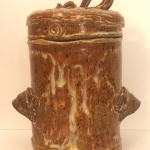 Peut inclure: Un pot en céramique brun avec un couvercle et deux poignées. Le pot a un glaçage moucheté avec des stries blanches et des taches brunes. Le couvercle a une poignée en boucle.