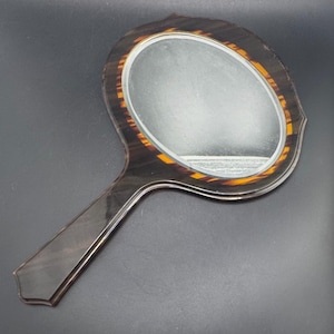 May include: A handheld mirror featuring an oval reflective surface framed by a dark brown and amber tortoiseshell pattern. The handle is long, straight, and made of a smooth, dark brown material that matches the outer edge of the mirror frame.
