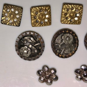 May include: A collection of vintage metal buttons in a variety of shapes and designs. The buttons are silver and gold with intricate details. Some buttons feature a floral design, while others have a geometric design.