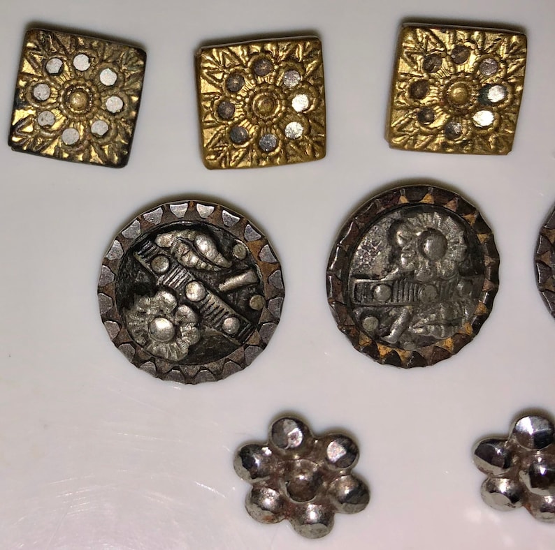 May include: A collection of vintage metal buttons in a variety of shapes and designs. The buttons are silver and gold with intricate details. Some buttons feature a floral design, while others have a geometric design.