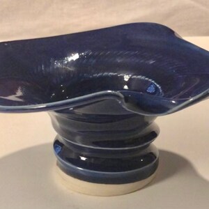 May include: A handcrafted, dark blue ceramic bowl with a unique, ruffled rim. The bowl sits atop a spiraled, cylindrical base, and a white base. This decorative piece is ideal for displaying small items or as a standalone art object.