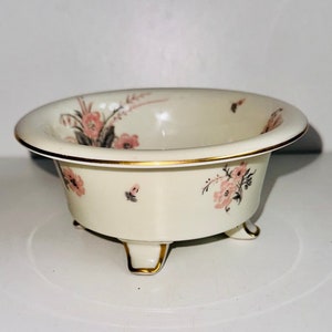 Può includere: Ciotola color crema con disegni floreali rosa e bordi dorati. La ciotola ha una forma arrotondata con un'ampia apertura e tre piedini. Il motivo floreale comprende fiori rosa e dettagli grigi.