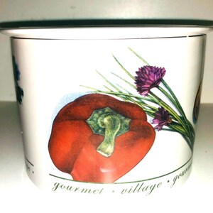 May include: White ceramic jar with a lid, featuring a red bell pepper, green chives, and purple flowers. The jar has the text "gourmet village gourmet" printed on the bottom.