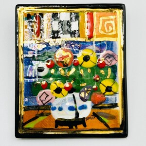 May include: A colorful ceramic tile with a black frame and gold accents. The tile features a painting of a vase with flowers in a variety of colors, including red, yellow, and blue. The tile is signed "Tommi" in the lower right corner.