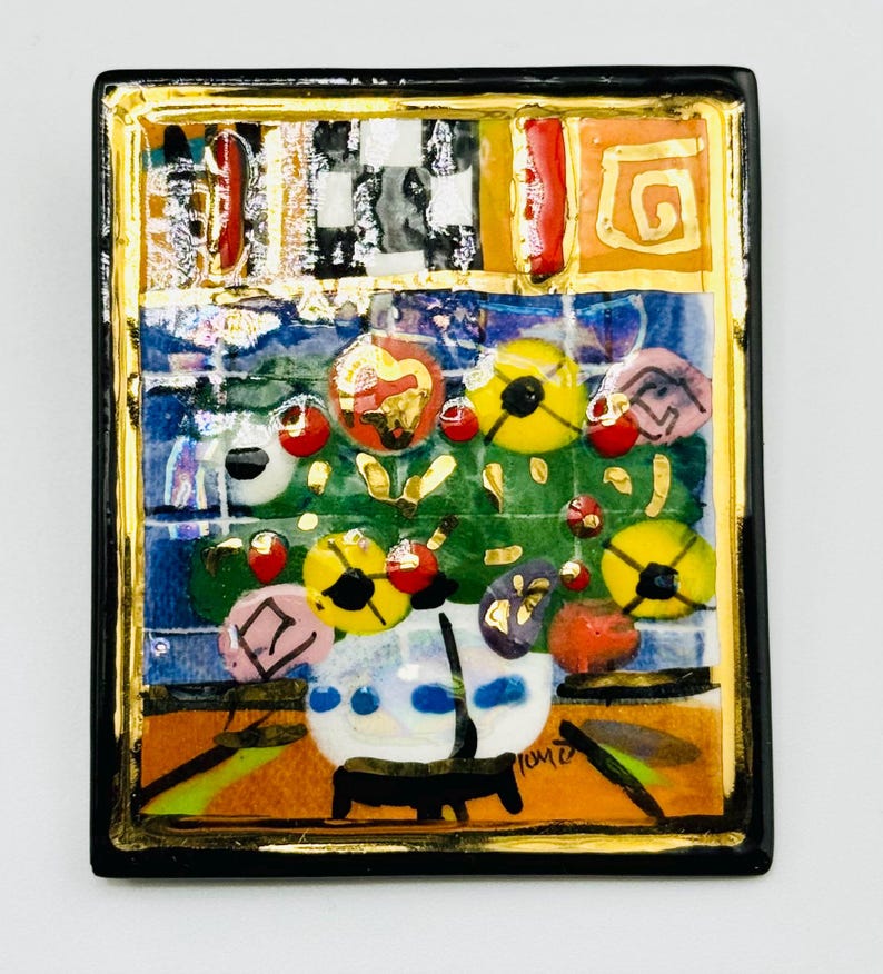 May include: A colorful ceramic tile with a black frame and gold accents. The tile features a painting of a vase with flowers in a variety of colors, including red, yellow, and blue. The tile is signed "Tommi" in the lower right corner.